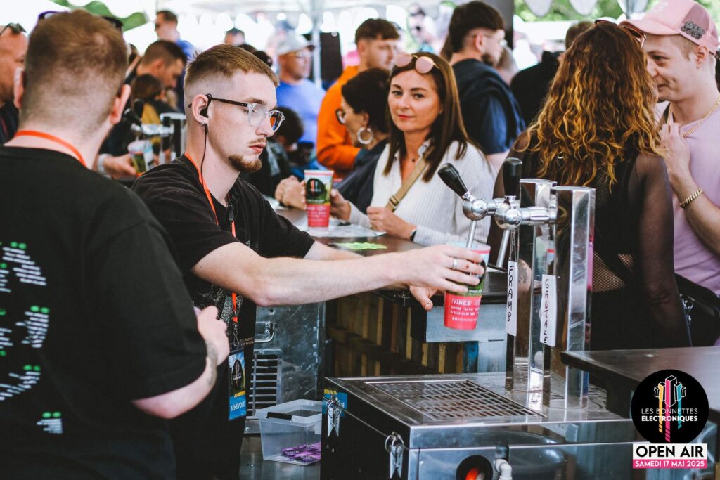 Bénévole du festival Les Bonnettes Électroniques servant des boissons au bar lors de l'édition 2025 à Étaing.